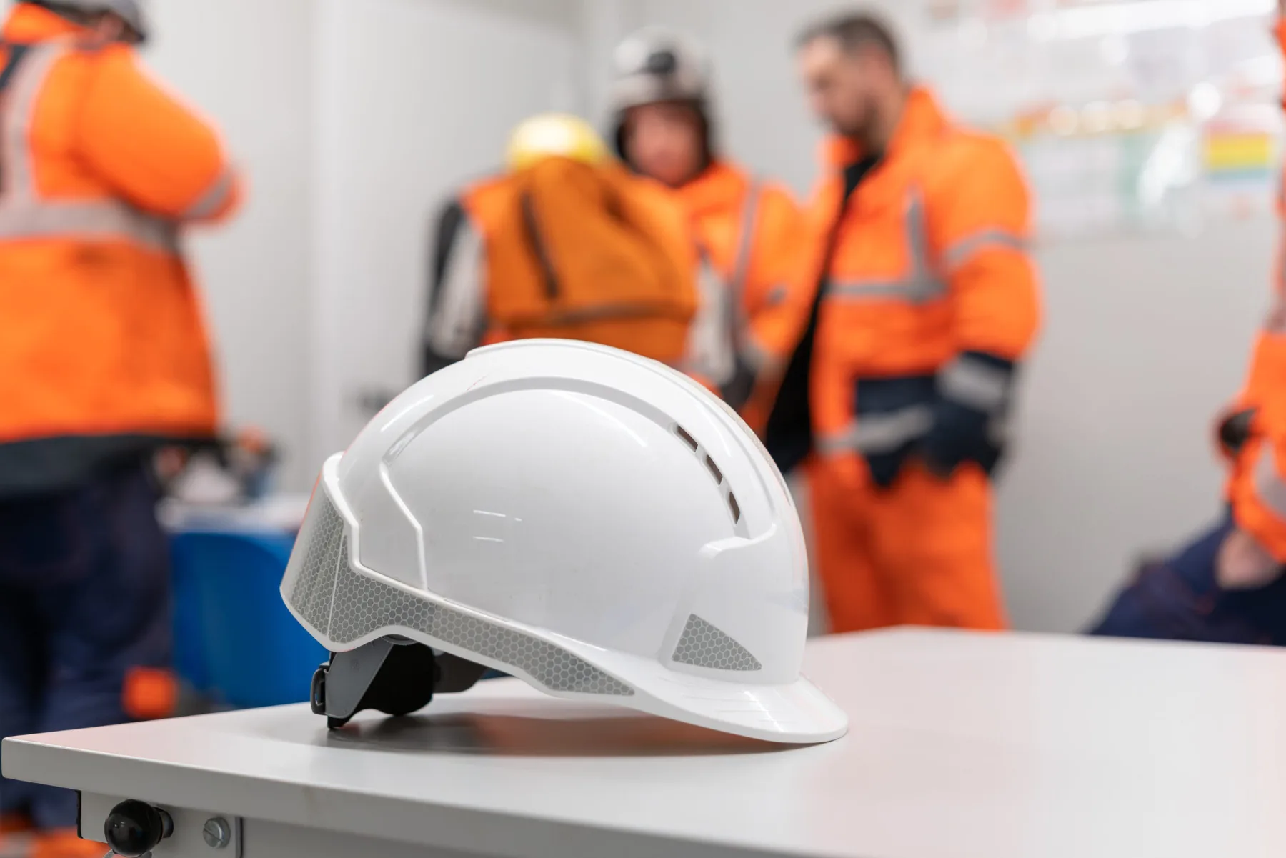 Casque de sécurité blanc posé sur une table, ouvriers en orange en arrière-plan
